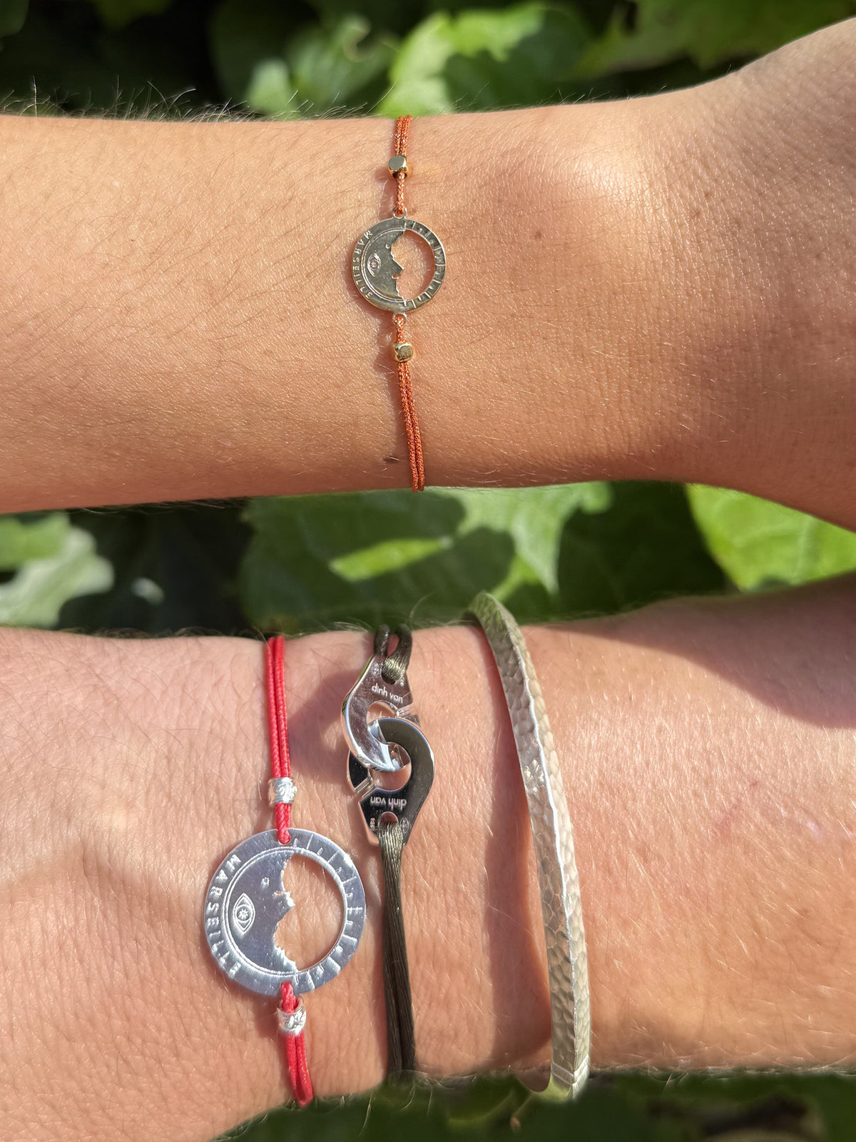 bracelets Ligne Bleue portés sur deux poignets : grande médaille Marseille argentée sur cordon rouge et petite médaille argentée sur cordon orangé, en plein soleil — Ligne Bleue, Marseille